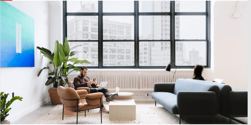 Waiting room with modern furniture with man sitting on chair, looking at phone and laptop, with cityscape view outside of window.
