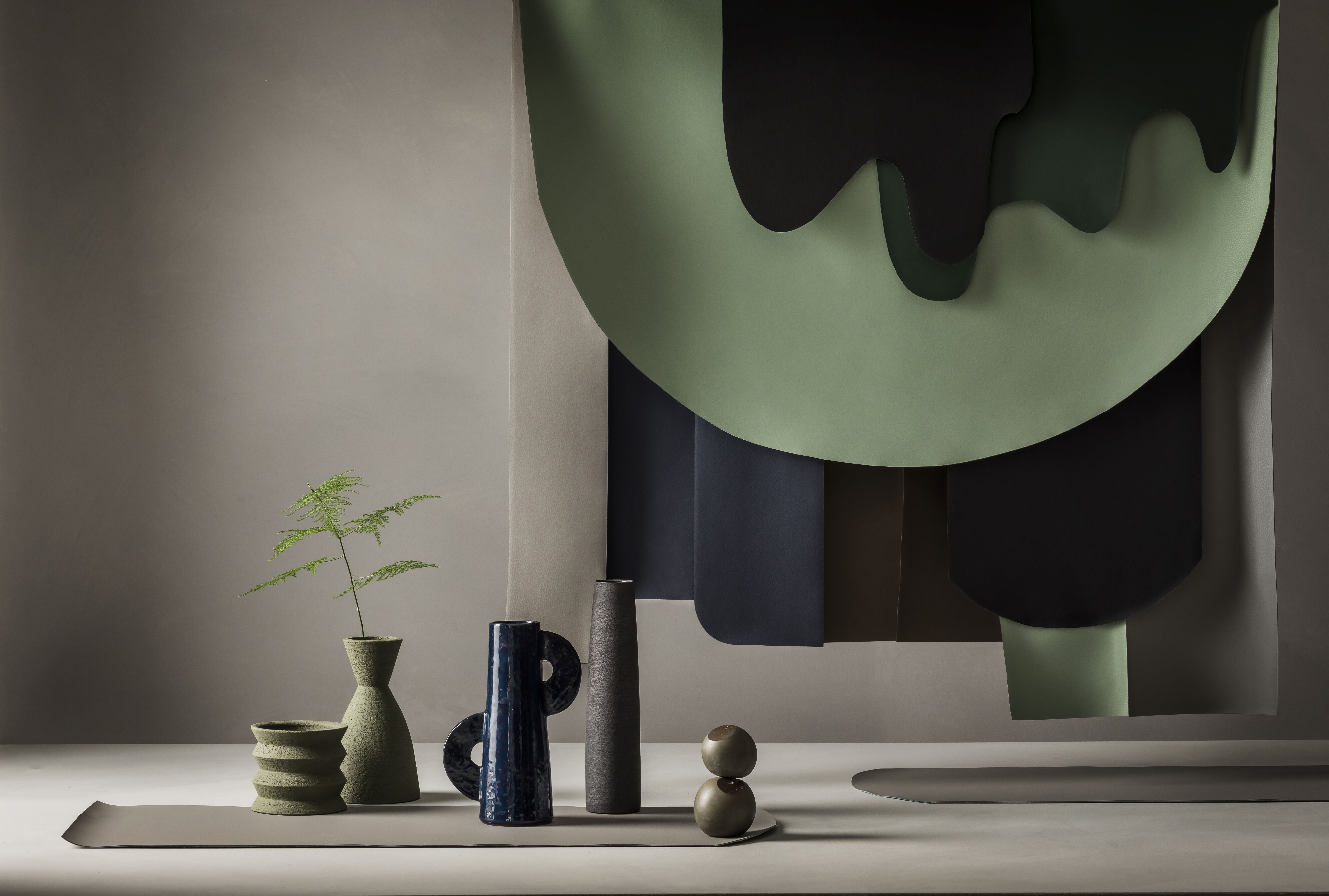 Still life with green and blue colored ceramics in the foreground and dark green, light green, and dark blue draped fabric in the background.