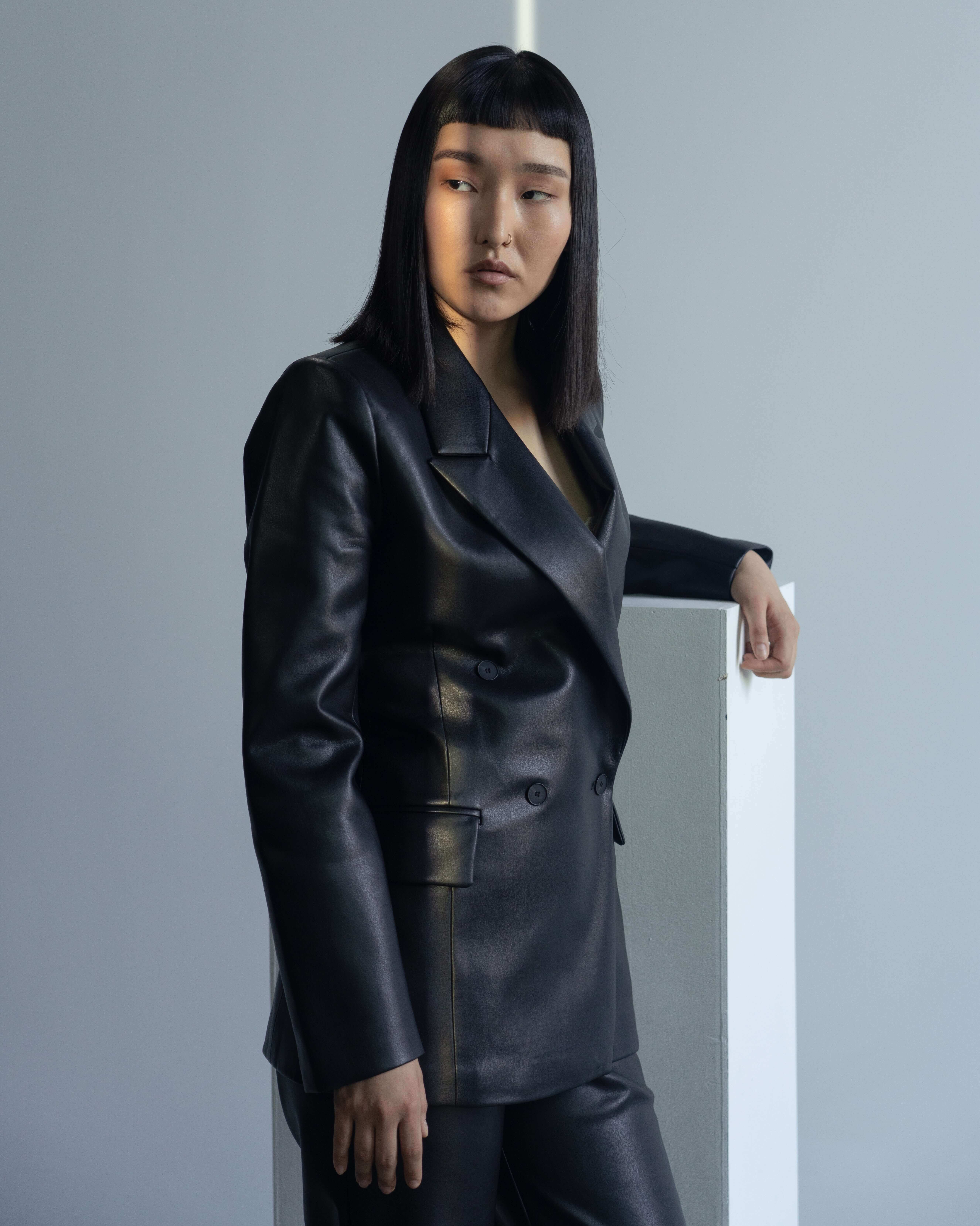 Woman model with dark, straight, shoulder-length hair, in black leather blazer and pants, resting arm on a white pedestal, looking over her shoulder.