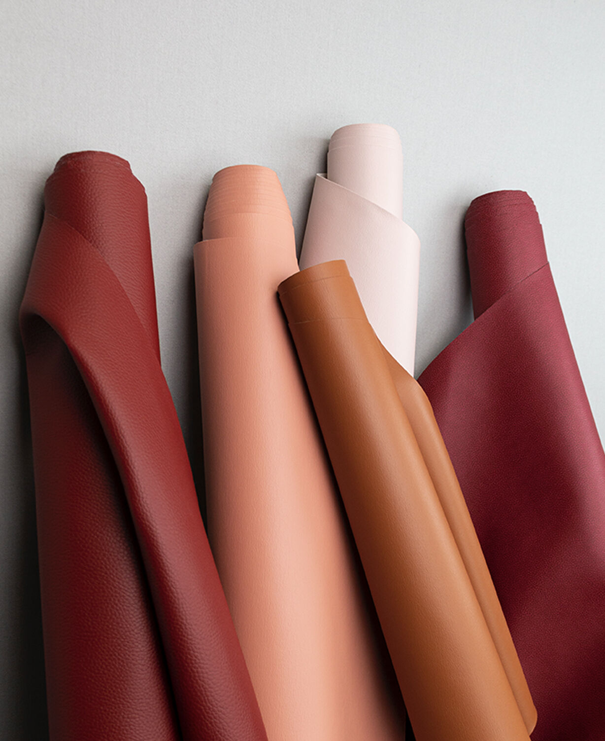 A red roll of fabric, a peach roll of fabric, an off-white roll of fabric, and a berry red roll of fabric lying next to each other with a tan roll of fabric on top.