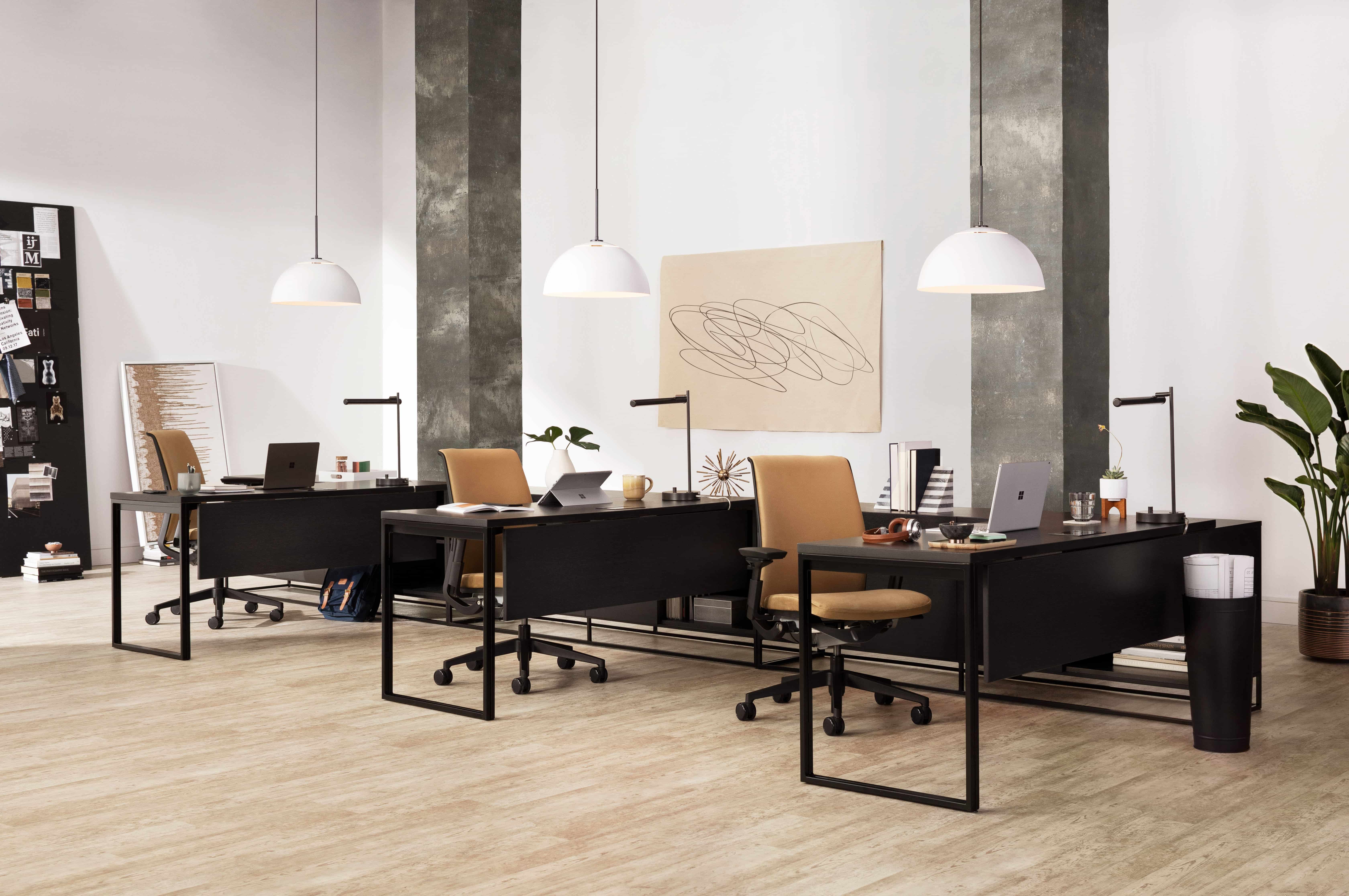 Modern office with black desks, tan swivel chairs, hanging white lights, and artwork on walls.